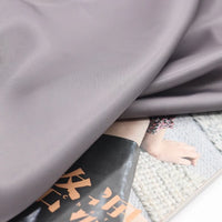 A swatch of Longan Craft’s Colorful Satin Viscose Fabric with twill weave in light gray drapes over a magazine page, partially obscuring an image of legs on a rug, with Japanese text visible at the bottom.