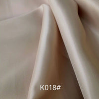 A close-up shows the silky, light beige Colorful Satin Viscose Fabric by Longan Craft with gentle folds and a slight sheen. “K018#” is displayed at the bottom center. This polyester viscose twill fabric is ideal for formal dresses.