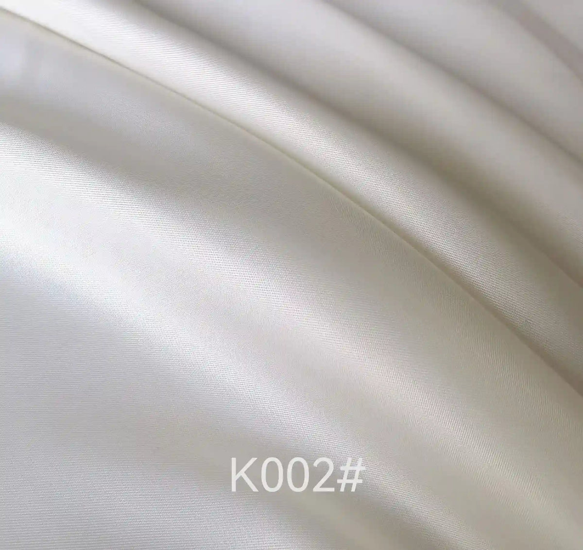 Close-up of smooth, shiny ivory or light cream Colorful Satin Viscose Fabric By The Yard from Longan Craft, featuring gentle folds and highlights. The text K002# is displayed at the bottom center of the image.