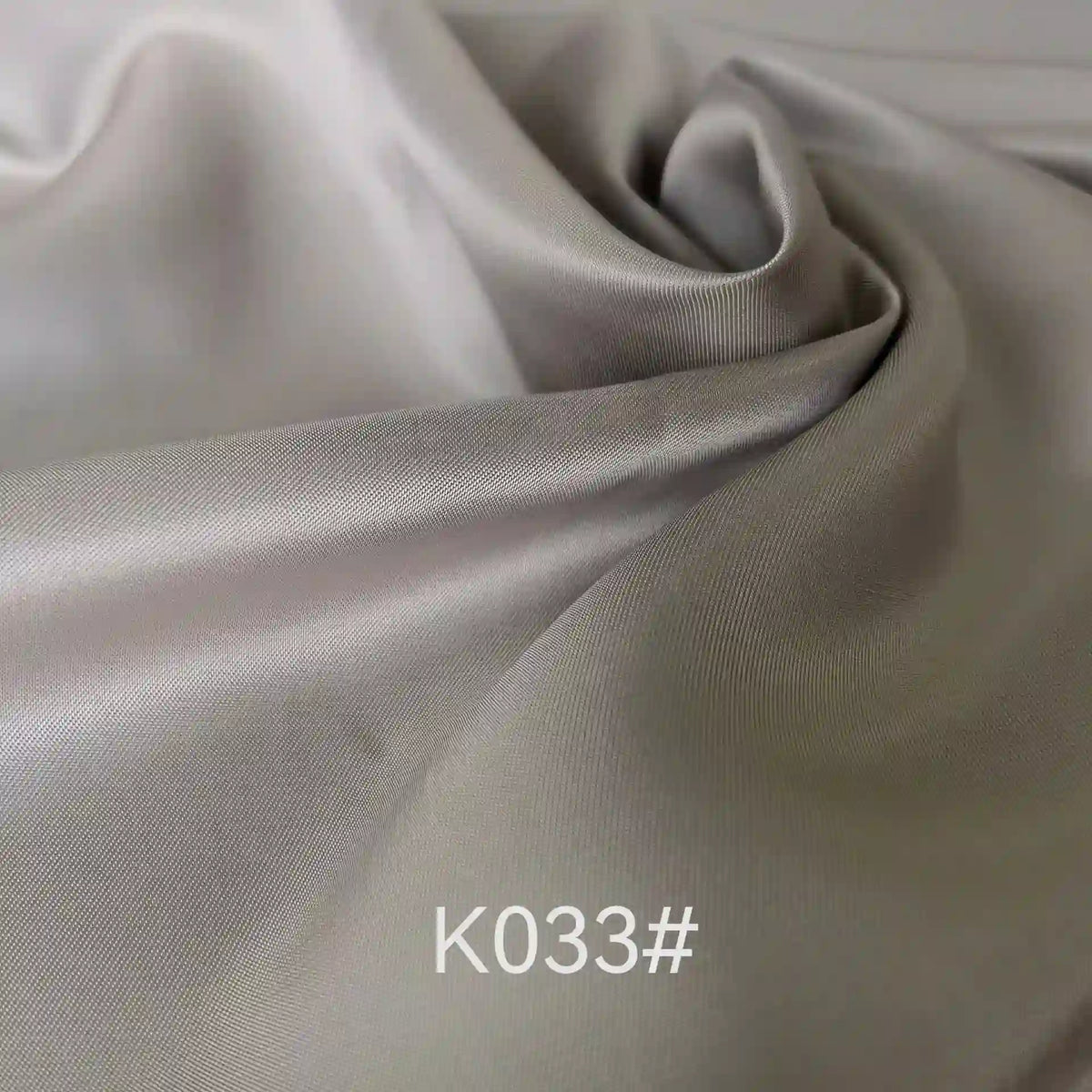 A close-up of elegant, light gray Colorful Satin Viscose Fabric by Longan Craft with gentle folds and a soft sheen. The text K033# appears in white at the bottom, showcasing this twill weave polyester viscose ideal for formal dresses.