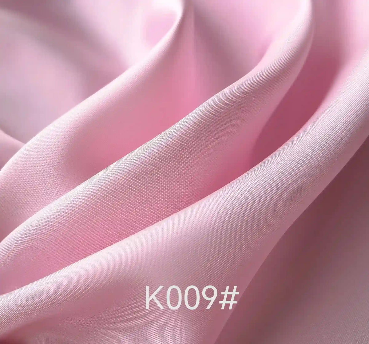 A close-up of Longan Craft Colorful Satin Viscose Fabric By The Yard with a twill weave shows smooth, light pink folds and a satin-like texture. The code K009# appears at the bottom center. Ideal for elegant and formal dresses.