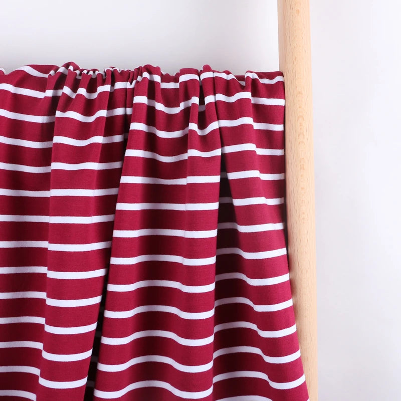A piece of Longan Craft Colorful Striped Stretch French Terry Fabric, a knit cotton French terry material ideal for sweatshirts, in maroon with white horizontal stripes is draped over a wooden rod against a plain white background.