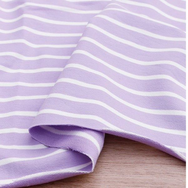 A piece of Longan Craft Colorful Striped Stretch French Terry Fabric by the yard in lavender and white is neatly folded on a wooden surface. This knit cotton French terry material is perfect for making sweatshirts.