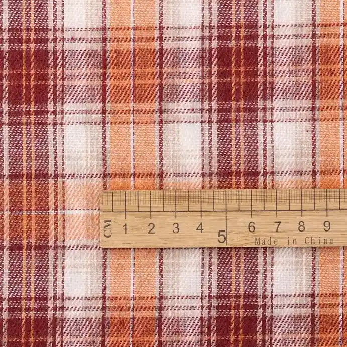 A wooden ruler marked with both centimeters and inches, and stamped with “Made in China,” is placed on Longan Craft’s Cotton Jacquard Plaid Flannel Fabric By The Yard. The cozy autumn flannel fabric features a red, orange, and white checked plaid pattern with intersecting lines and squares, characteristic of one-sided brushed flannel material ideal for clothing.