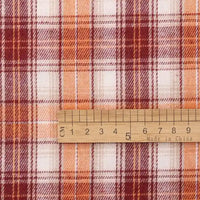 A wooden ruler marked with both centimeters and inches, and stamped with “Made in China,” is placed on Longan Craft’s Cotton Jacquard Plaid Flannel Fabric By The Yard. The cozy autumn flannel fabric features a red, orange, and white checked plaid pattern with intersecting lines and squares, characteristic of one-sided brushed flannel material ideal for clothing.