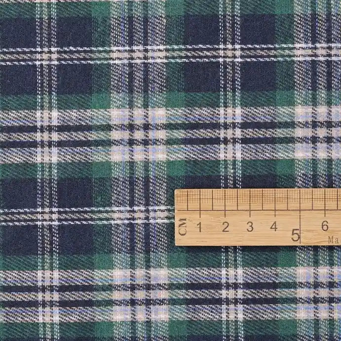 A wooden ruler marked in centimeters provides a sense of scale for the green, navy, and white plaid pattern on the Cotton Jacquard Plaid Flannel Fabric by the yard from Longan Craft. This one sided brushed checked flannel material is perfect for cozy autumn clothing projects.