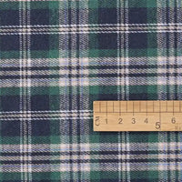 A wooden ruler marked in centimeters provides a sense of scale for the green, navy, and white plaid pattern on the Cotton Jacquard Plaid Flannel Fabric by the yard from Longan Craft. This one sided brushed checked flannel material is perfect for cozy autumn clothing projects.