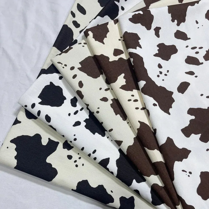 Multiple pieces of Longan Craft Cow Print Denim Fabric By The Yard (100% cotton denim) in black and brown cow-like splotches are neatly layered, showcasing varying pattern intensities—perfect DIY handmade clothing twill material.
