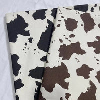 Two pieces of Longan Craft Cow Print Denim Fabric By The Yard, made from 100% cotton denim, are shown side by side: black spots on white and brown spots on white, both displayed on a light, textured background.