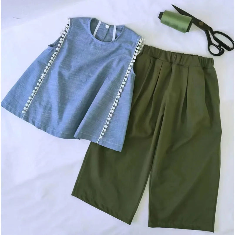 A blue sleeveless top with white trim is paired with high-waisted green pants made using the Longan Craft Wide Leg Capri Pant Sewing Pattern, shown beside a spool of green thread and black scissors on a white surface.