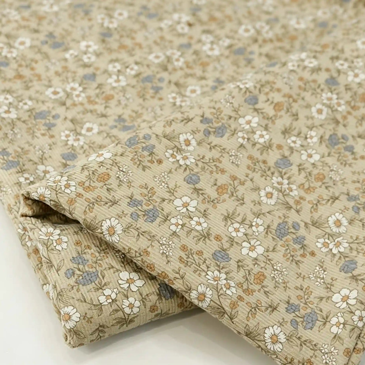 The Longan Craft Daisy Print 16 Wale Corduroy Fabric By The Yard features a delicate beige base adorned with small white daisies, blue and tan flowers, and green leaves. This fine wale 100% cotton corduroy material displays a subtle textured pattern and is shown folded on a white surface—perfect for fall DIY projects.