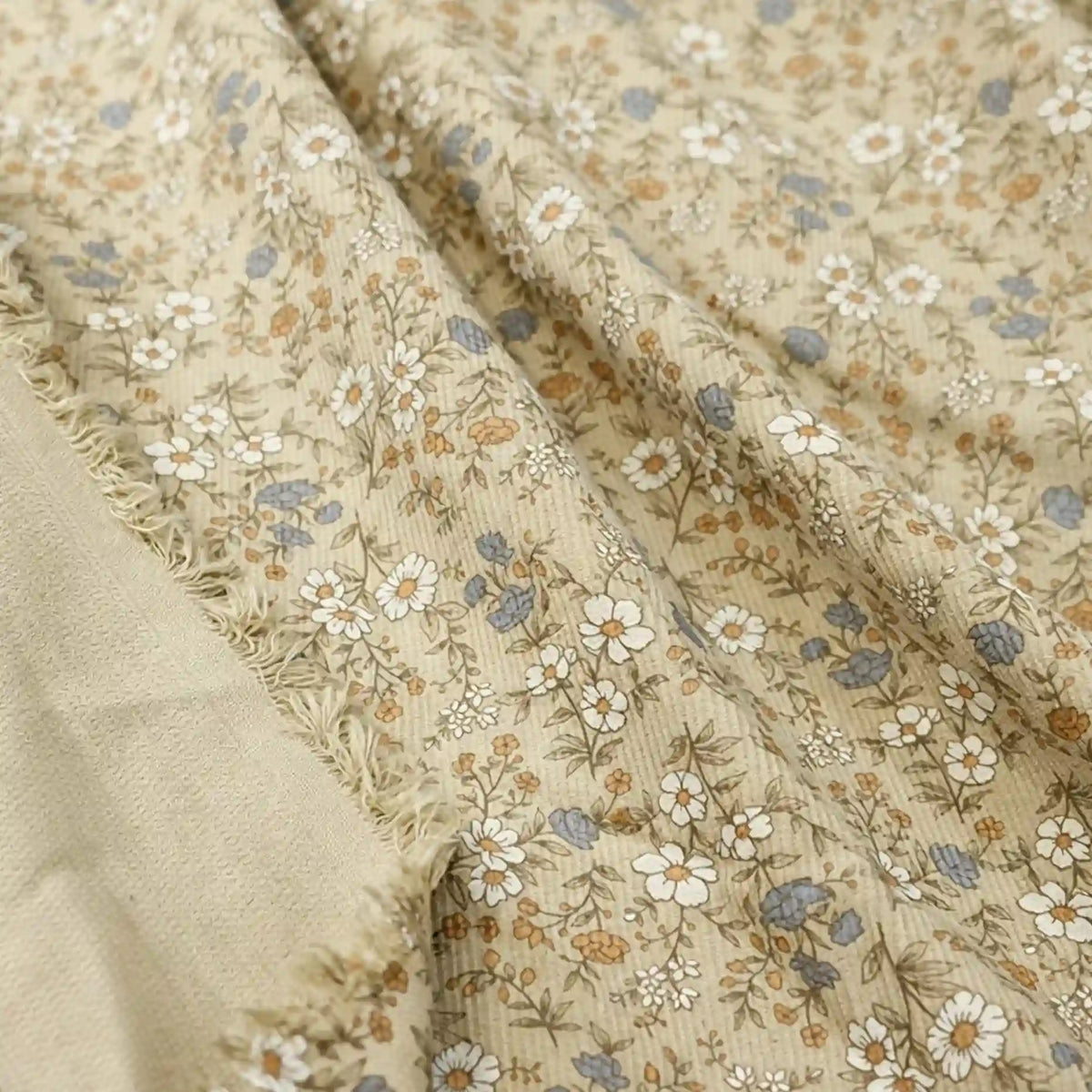 Longan Craft’s Daisy Print 16 Wale Corduroy Fabric by the yard features a fine wale, 100% cotton corduroy material in beige with a delicate floral design of white, blue, and brown flowers with green stems. This fall-inspired DIY fabric offers a soft, textured surface and has one edge slightly frayed to reveal the fabric’s underside.