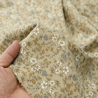 A hand holds a piece of the Daisy Print 16 Wale Corduroy Fabric by Longan Craft, featuring a fine beige corduroy with a delicate pattern of white, blue, and brown flowers with green leaves. This soft 100% cotton corduroy material is slightly wrinkled and perfect for adding a charming touch to any fall DIY project.