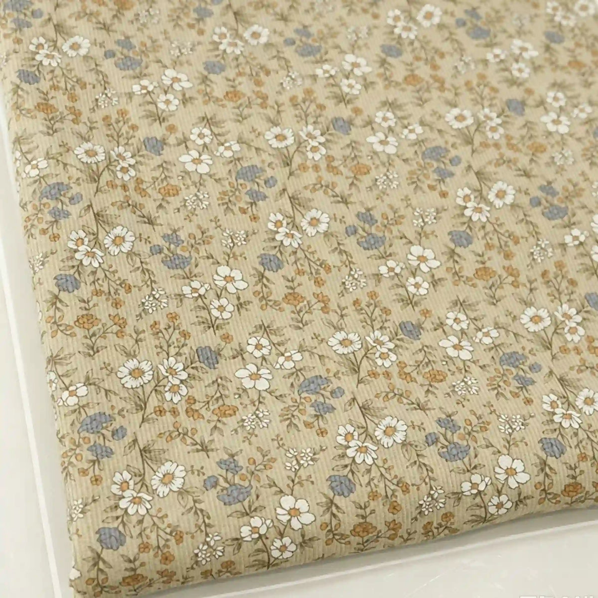 A folded piece of Longan Craft Daisy Print 16 Wale Corduroy Fabric, made from 100% fine wale cotton, displays a quilted beige surface adorned with white daisies, small blue flowers, and brown leaves—a perfect fall corduroy material for DIY projects.