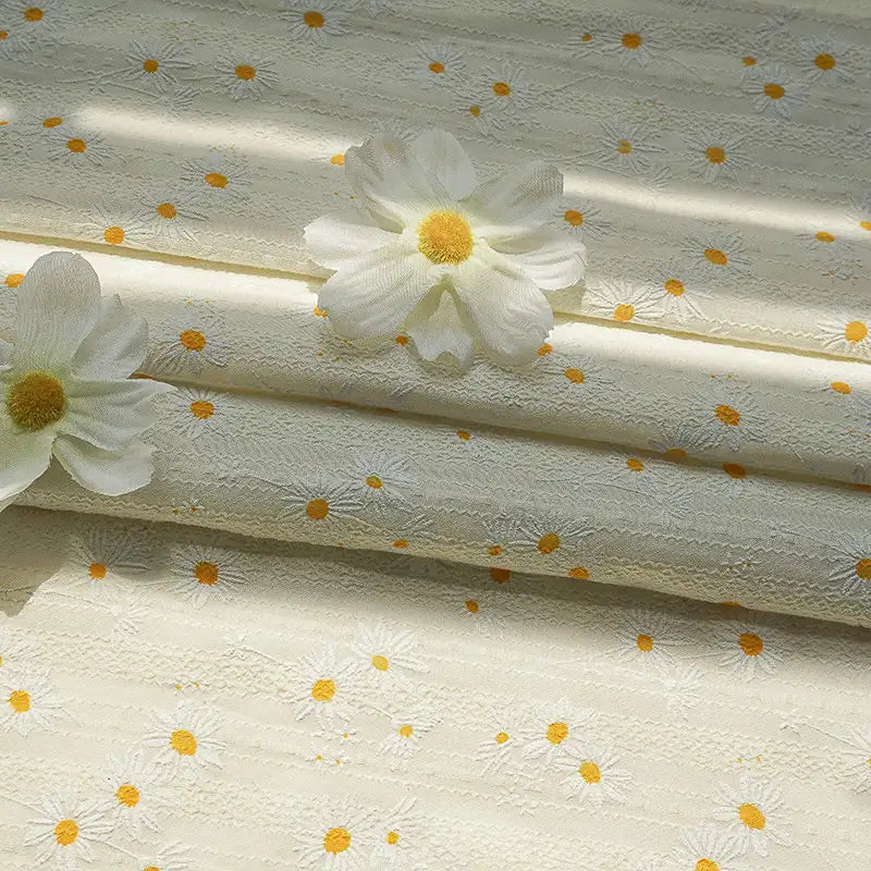 Longan Craft Daisy Printed Dobby Fabric By The Yard features subtle cream and yellow daisy patterns. This cozy, textured cotton material is ideal for clothing, shown gently folded and accented with two artificial daisies on top.
