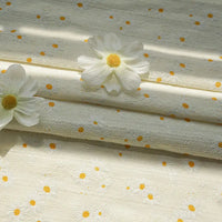 Longan Craft Daisy Printed Dobby Fabric By The Yard features subtle cream and yellow daisy patterns. This cozy, textured cotton material is ideal for clothing, shown gently folded and accented with two artificial daisies on top.