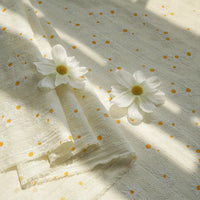 Longan Craft's Daisy Printed Dobby Fabric by the yard features small yellow and white daisy motifs on cream cotton textured dobby, displayed flat with two faux daisies as sunlight highlights the cozy, floral surface—perfect for clothing projects.