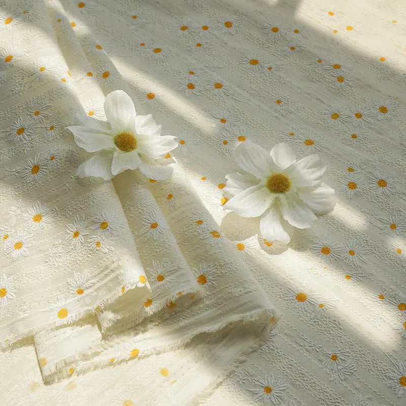 Longan Craft's Daisy Printed Dobby Fabric by the yard features small yellow and white daisy motifs on cream cotton textured dobby, displayed flat with two faux daisies as sunlight highlights the cozy, floral surface—perfect for clothing projects.