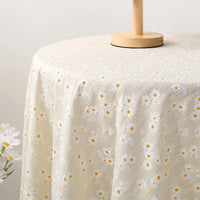 A round table is covered with Longan Craft’s Daisy Printed Dobby Fabric By The Yard, featuring cream-colored cotton adorned with small white daisies and yellow centers. A wooden stand sits on the fabric, with a vase of daisies in the background.