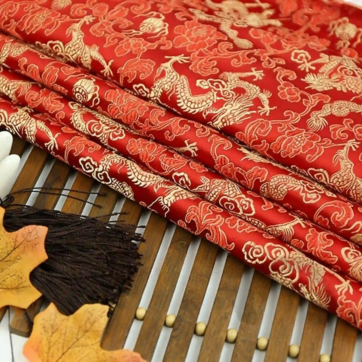 A stack of Dragon Pattern Brocade Fabric By The Yard by Longan Craft, featuring gold dragons and clouds, rests on a bamboo mat with decorative autumn leaves and a black tassel nearby. This multicolor vintage chinoiserie material adds elegance to any project.