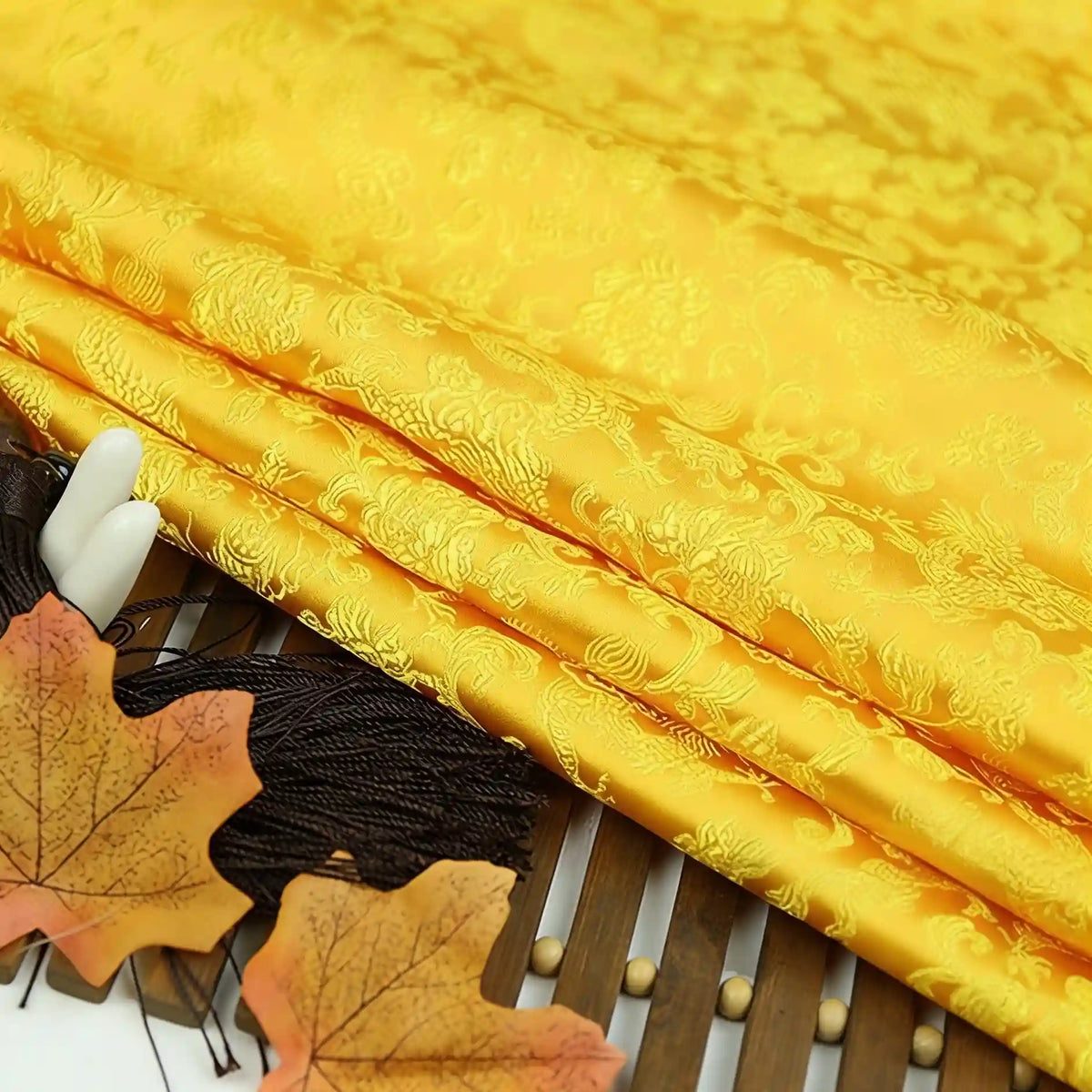 Longan Craft's Dragon Pattern Brocade Fabric By The Yard – a multicolor Chinoiserie vintage cloth with intricate patterns, shown folded on bamboo, accented by two autumn leaves and black tassels.