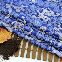 Longan Craft's Dragon Pattern Brocade Fabric By The Yard features multicolor chinoiserie dragon and floral designs. This vintage brocade material is shown folded on a wooden surface, accented by dried leaves and a dark brown fringe.