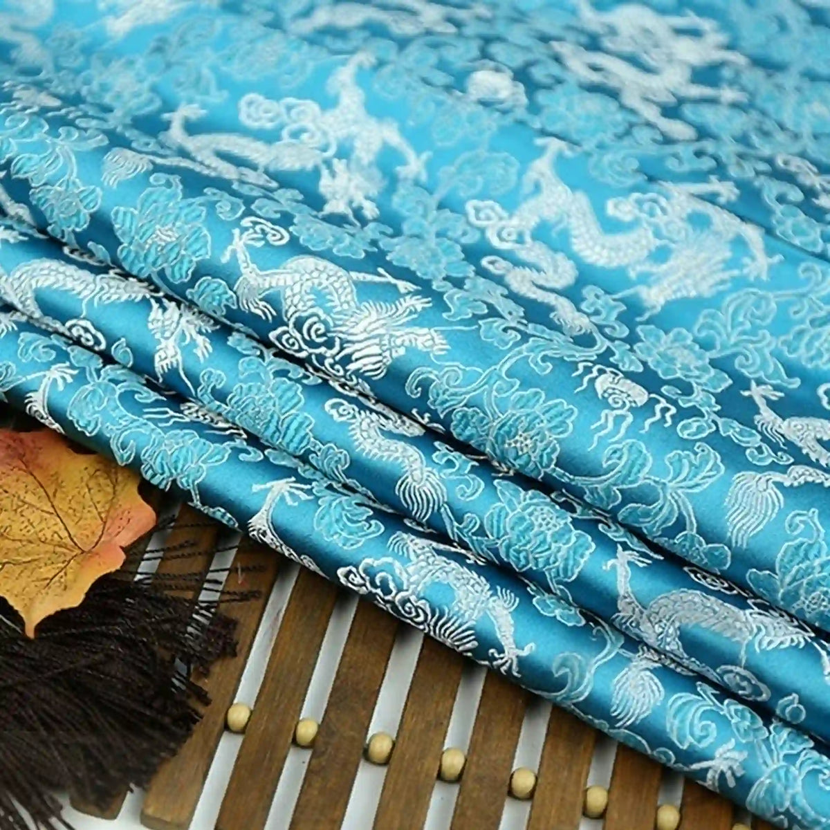 The Longan Craft Dragon Pattern Brocade Fabric by the yard showcases multicolor chinoiserie dragon and cloud motifs in blue, artfully displayed with black tassels and a decorative leaf on a wooden mat.