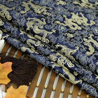 A close-up of Longan Craft's Dragon Pattern Brocade Fabric By The Yard, a multicolor Chinoiserie brocade material, featuring intricate gold dragons and clouds, folded on a bamboo mat with brown tassels and autumn leaves nearby.