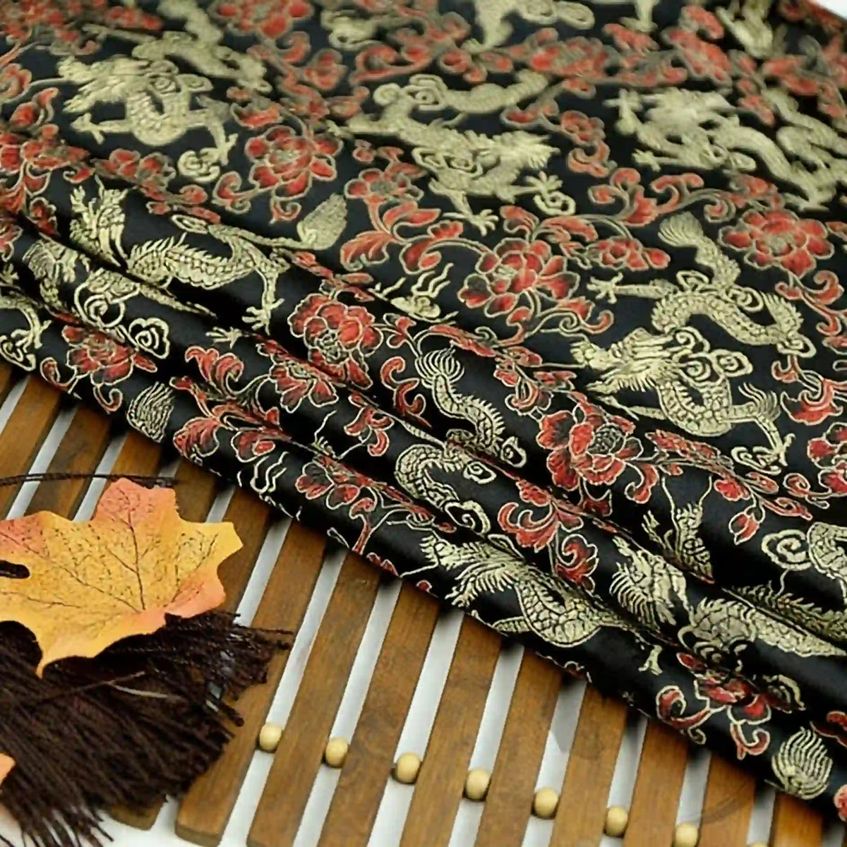 A close-up of Longan Craft’s Dragon Pattern Brocade Fabric By The Yard—multicolor chinoiserie material with intricate gold and red dragon motifs—shown folded on a bamboo mat with autumn leaves and a brown tassel nearby.