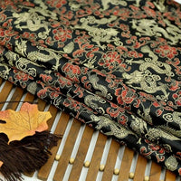 A close-up of Longan Craft’s Dragon Pattern Brocade Fabric By The Yard—multicolor chinoiserie material with intricate gold and red dragon motifs—shown folded on a bamboo mat with autumn leaves and a brown tassel nearby.