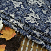 A stack of Longan Craft Dragon Pattern Brocade Fabric By The Yard, a multicolor vintage Chinoiserie material with white dragon and cloud motifs, is displayed on wood beside brown fringe and autumn leaves.