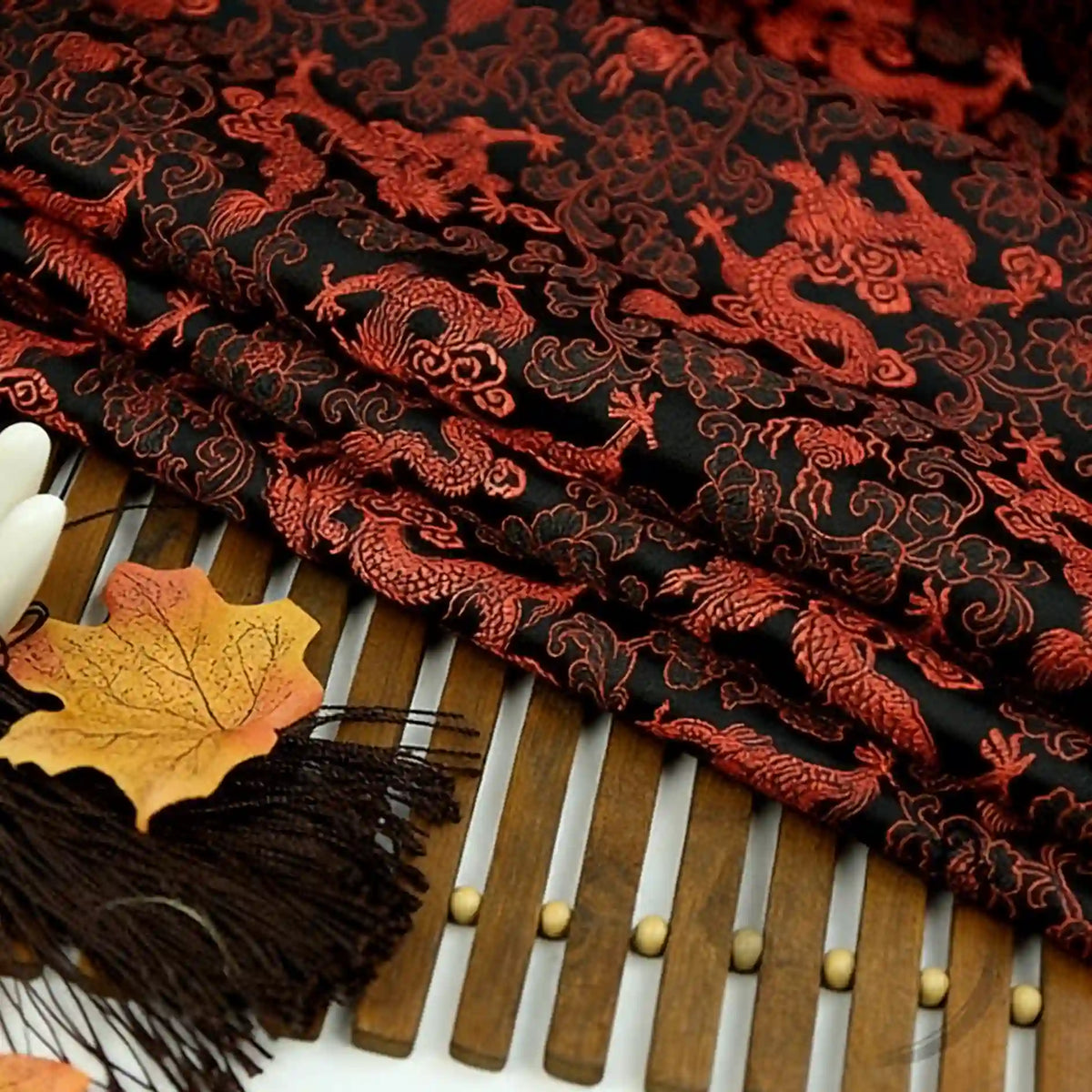 Longan Craft's Dragon Pattern Brocade Fabric By The Yard, a multicolor chinoiserie brocade cloth, is elegantly folded on a bamboo mat with autumn leaves and a dark brown tassel beside it.