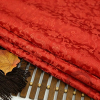 The Dragon Pattern Brocade Fabric by Longan Craft, featuring vibrant multicolor chinoiserie designs, is displayed folded on a wooden surface beside a brown tassel and an autumn leaf.