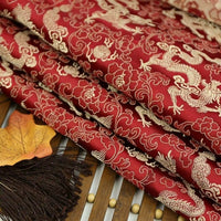 Longan Craft Dragon Pattern Brocade Fabric by the yard features intricate gold dragons and floral motifs in multicolor chinoiserie style, shown folded on a wooden surface beside a decorative fan and an artificial autumn leaf.