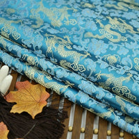 Longan Craft's Dragon Pattern Brocade Fabric By The Yard, a multicolor vintage chinoiserie brocade material with gold dragon and floral designs, is displayed folded on wood with autumn leaves, brown fringe, and white decorative sticks.