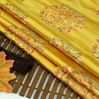 A close-up of Longan Craft's Dragon Pattern Brocade Fabric by the yard, featuring multicolor chinoiserie embroidery, displayed on a bamboo mat next to a brown tassel and an autumn leaf.