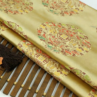 The Longan Craft Dragon Pattern Brocade Fabric By The Yard features intricate circular motifs in red, green, and gold tones. This vintage multicolor chinoiserie brocade material is displayed on a wooden mat with a dark brown tassel nearby.