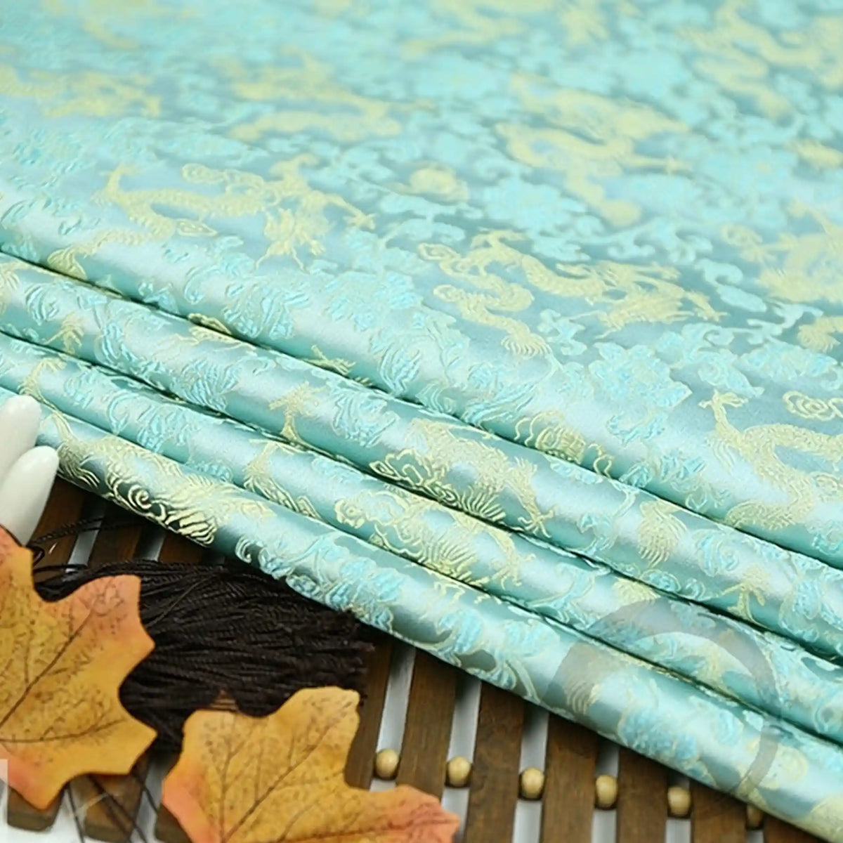 Longan Craft's Dragon Pattern Brocade Fabric by the yard, a multicolor Chinoiserie material with gold dragon motifs, is displayed on a bamboo mat and accented with artificial autumn leaves in the foreground.