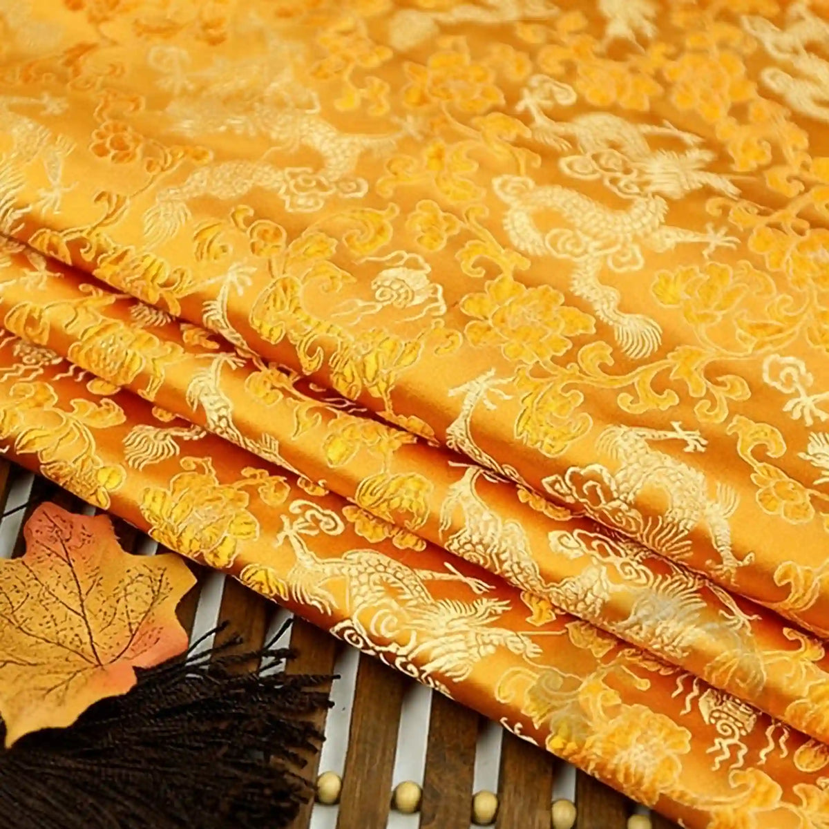 A close-up of Longan Craft’s Dragon Pattern Brocade Fabric By The Yard, a multicolor chinoiserie vintage brocade cloth with dragon and floral motifs, shown folded on a bamboo mat with a leaf and black tassel beside it.