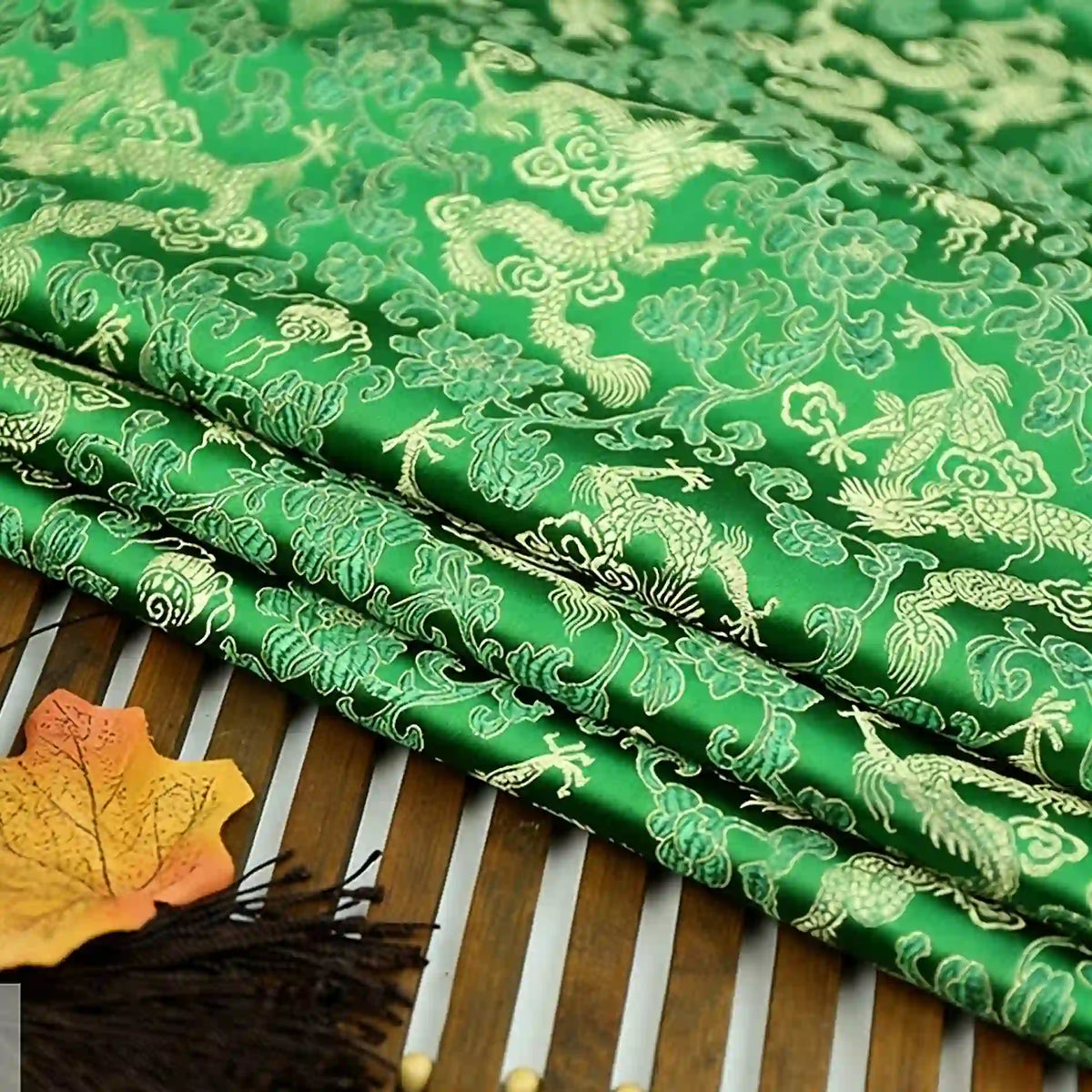 The Dragon Pattern Brocade Fabric By The Yard from Longan Craft features multicolor chinoiserie designs with gold dragons and clouds. This vintage brocade material is displayed folded on a bamboo mat, accented by an orange-yellow artificial leaf and fringe.