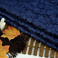 A piece of Longan Craft Dragon Pattern Brocade Fabric in multicolor Chinoiserie style is folded on a wooden mat, accented with artificial autumn leaves and black tassels for decoration.