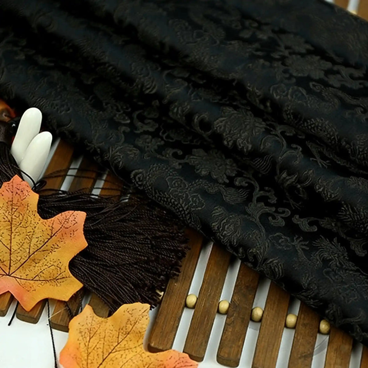 A piece of Longan Craft Dragon Pattern Brocade Fabric By The Yard, featuring a vintage multicolor chinoiserie design, is displayed on a wooden surface with artificial autumn leaves and brown tassels.