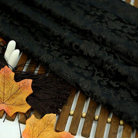 A piece of Longan Craft Dragon Pattern Brocade Fabric By The Yard, featuring a vintage multicolor chinoiserie design, is displayed on a wooden surface with artificial autumn leaves and brown tassels.
