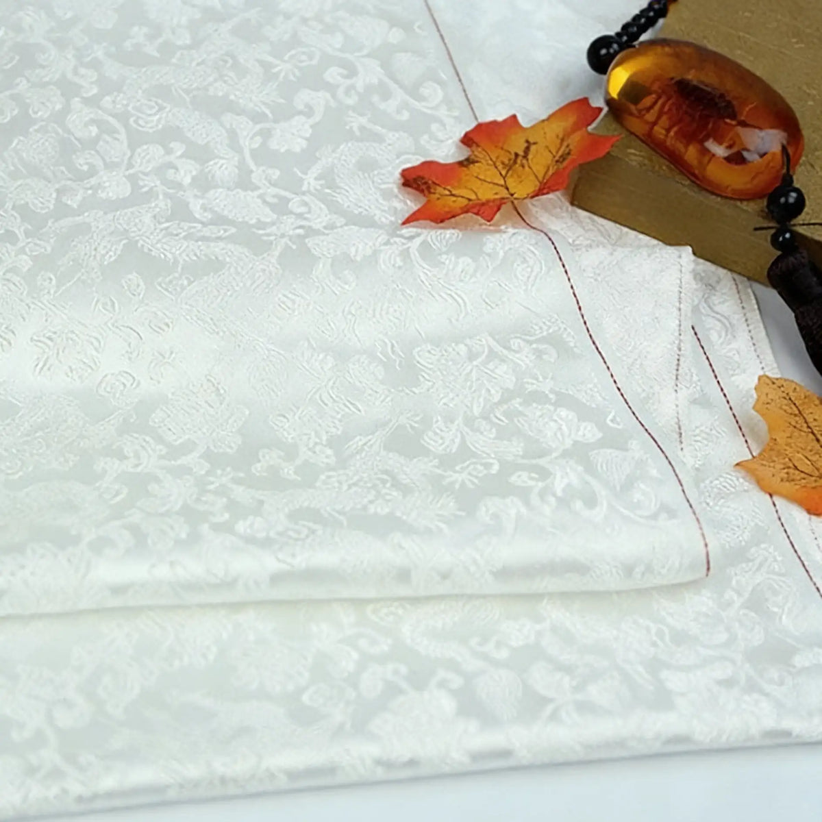 A piece of Longan Craft's Dragon Pattern Brocade Fabric by the Yard, a multicolor chinoiserie brocade with an intricate floral dragon motif, accented by orange autumn leaves and a beaded amber pendant on the side.