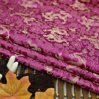 The Longan Craft Dragon Pattern Brocade Fabric By The Yard features multicolor chinoiserie dragon and floral designs. Folded on a bamboo mat, it's displayed with a decorative hairpin and an autumn leaf in the foreground.
