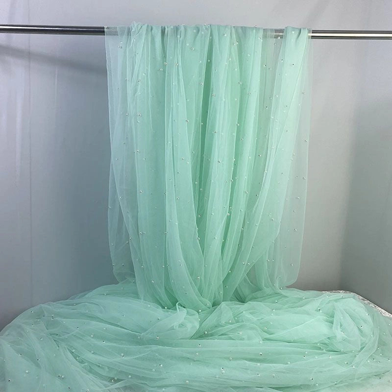 Longan Craft's Elegant Pearl Embellished Tulle Fabric By The Yard in mint green drapes softly over a rod, its sheer, lightweight material pooling below against a plain background.