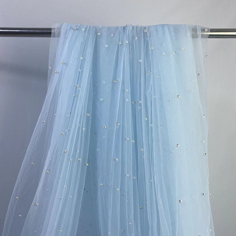 The Elegant Pearl Embellished Tulle Fabric by Longan Craft features sheer, light blue tulle adorned with evenly spaced pearl-like beads, draped over a metal rod and set against a plain light gray background.