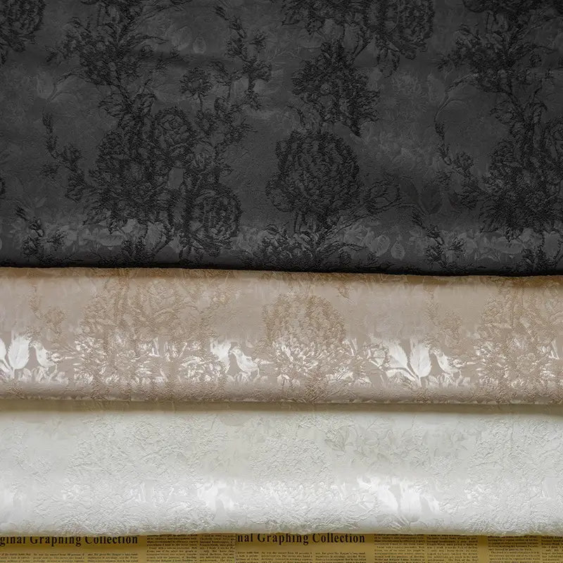 Three rolls of Longan Craft Embossed Peony Brocade Fabric in black, beige, and white are stacked horizontally on vintage newspaper print. Ideal Chinese-style material for dresses.