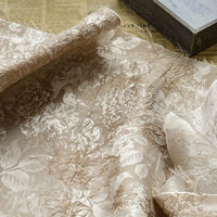 A close-up shows Longan Craft’s Embossed Peony Brocade Fabric By The Yard, a Chinese-style floral material for dresses, spread over an old newspaper with a closed book visible in the upper right corner.