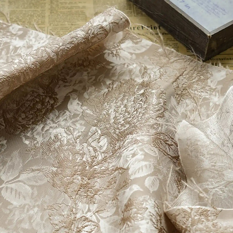 A close-up shows Longan Craft’s Embossed Peony Brocade Fabric By The Yard, a Chinese-style floral material for dresses, spread over an old newspaper with a closed book visible in the upper right corner.
